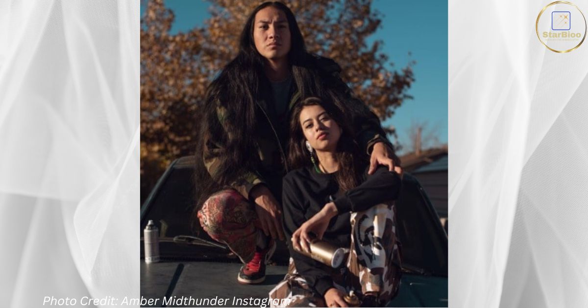 Amber Midthunder and her boyfriend River Thomas are sitting on a car. Midthunder is wearing a black sweatshirt and check cargo trousers, while Thomas is wearing a check jacket and pants. The couple seems happy and in love