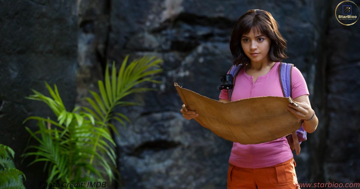 Isabela Merced in a still of Dora and the Lost City of Gold. She is wearing a pink tshirt and orange short with a map in her hand